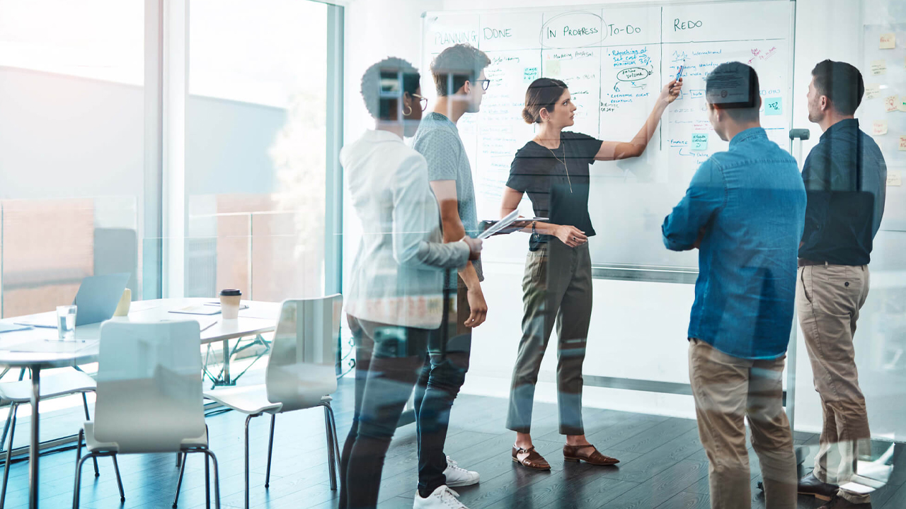 Fünf Personen stehen an Whiteboard und besprechen sich